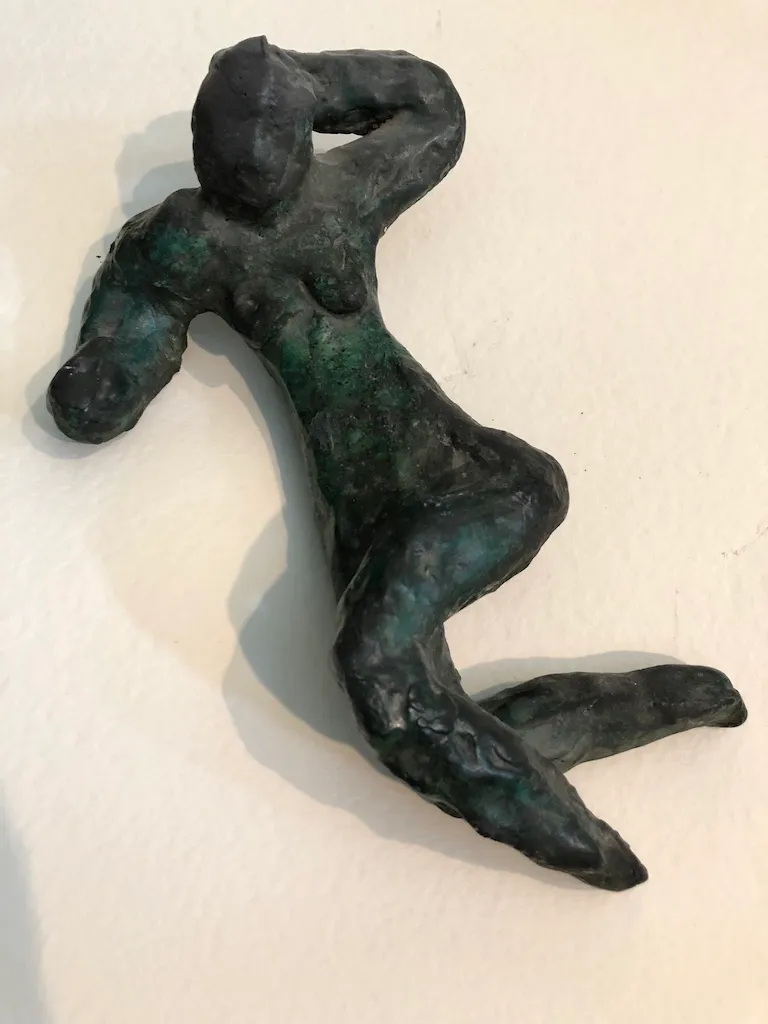 A small figure with a green-black patina, reclining with one arm folded behind her head and her legs drawn up.