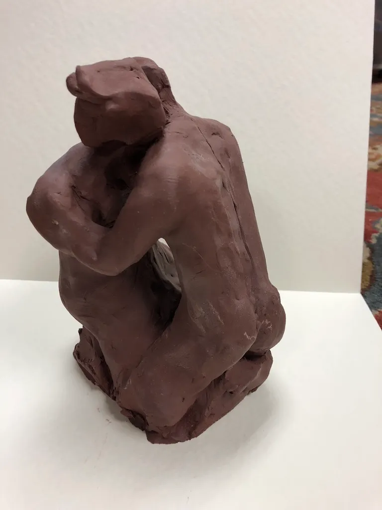 A red-brown clay figure kneeling on a rough base, its back curved and its arms crossed at its chest.