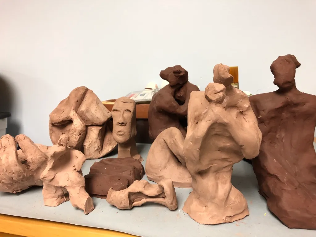 A studio table crowded with unglazed terracotta pieces — heads, torsos and an embracing pair grouped together.