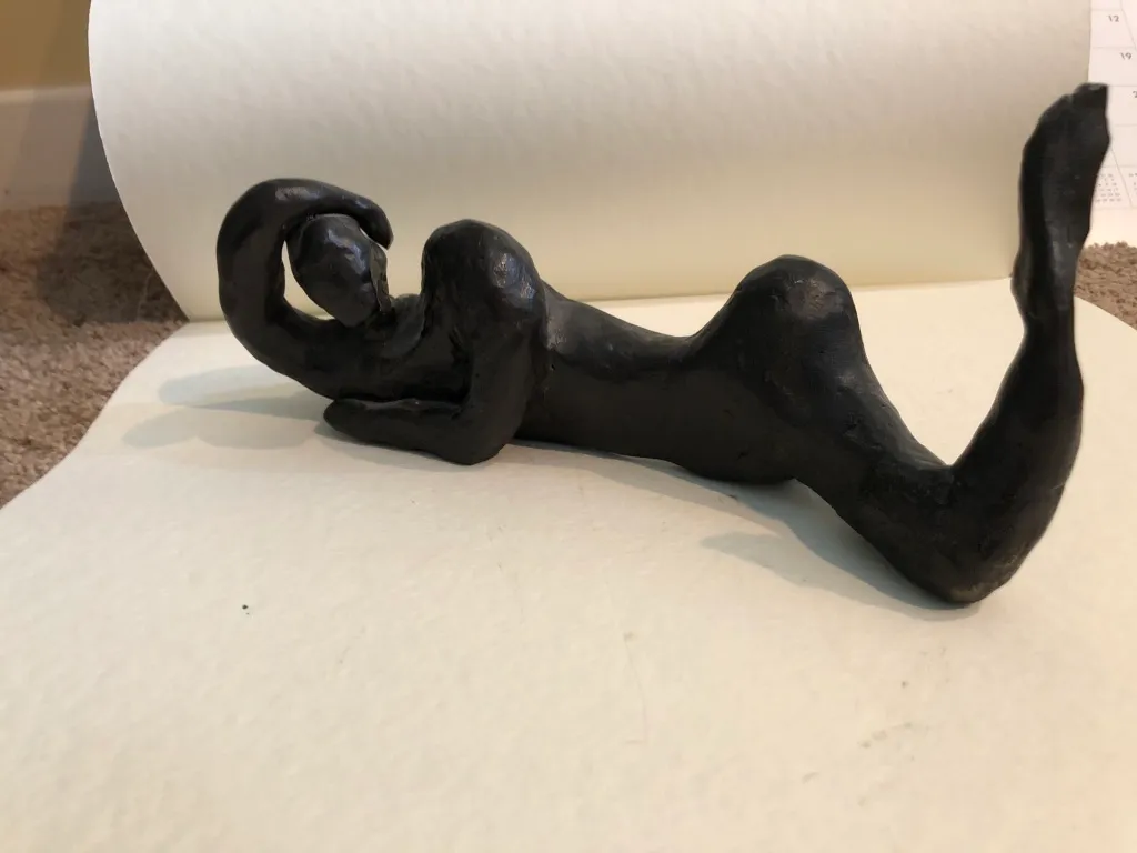 A black clay figure reclining on a sheet of paper, one leg lifted.