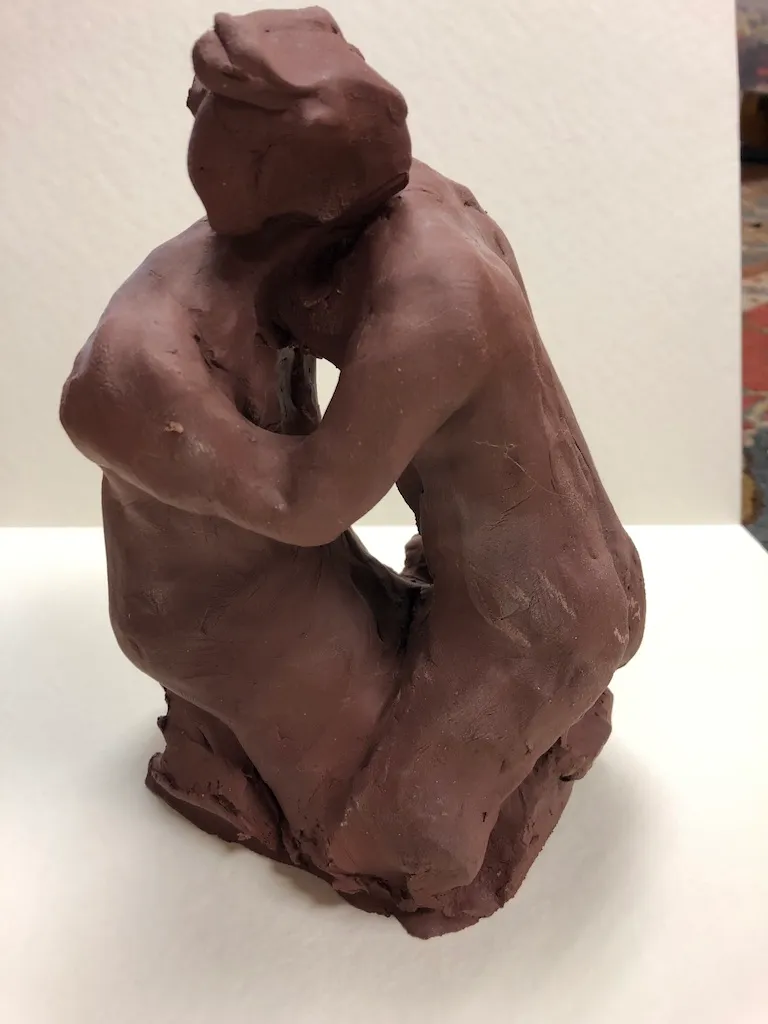 The kneeling clay figure from the side, its head lowered onto its folded arms.